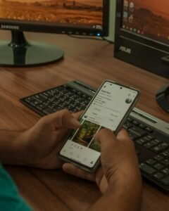 Person using a smartphone for digital marketing while working at a computer desk with multiple monitors.